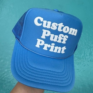 May include: A blue trucker hat with a white mesh back. The hat has white puff print lettering that says "Custom Puff Print".