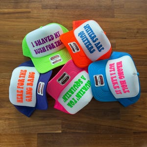 May include: Five foam trucker hats with mesh backs in neon green, orange, pink, blue, and purple. Each hat features a white front panel with printed text: I SHAVED MY BUSH FOR THIS, SPITTERS ARE QUITTERS, HERE FOR THE GANG BANG, SQUIRTIN' FOR A SQUIRTIN', and WRONG HOLE BUT I LIKE IT.