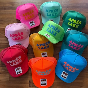 May include: A collection of colorful trucker hats with the text "APR&Egrave;S LAKE" in various colors. The hats are in shades of pink, green, blue, yellow, orange, and red. The hats have a mesh back and a curved brim.