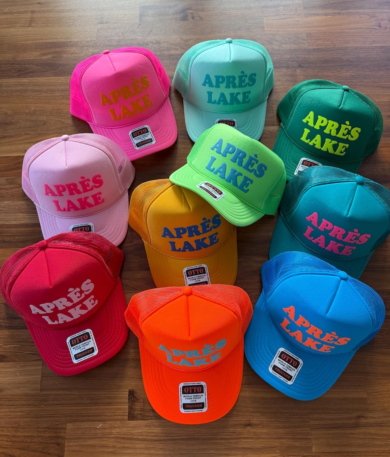 May include: A collection of colorful trucker hats with the text "APR&Egrave;S LAKE" in various colors. The hats are in shades of pink, green, blue, yellow, orange, and red. The hats have a mesh back and a curved brim.