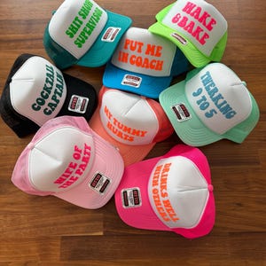 May include: A collection of seven foam and mesh trucker hats in various colors, each featuring a white front panel with bold text including SHIT SHOW SUPERVISOR, HAKE & BAKE, PUT ME IN COACH, COCKTAIL CAPTAIN, TWERKING 9 TO 5, MY TUMMY HURTS, WIFE OF THE PARTY, and DRINKS WELL WITH OTHERS.