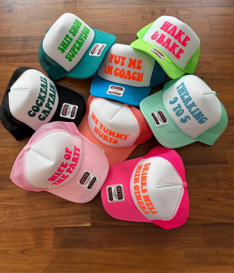 May include: A collection of seven foam and mesh trucker hats in various colors, each featuring a white front panel with bold text including SHIT SHOW SUPERVISOR, HAKE & BAKE, PUT ME IN COACH, COCKTAIL CAPTAIN, TWERKING 9 TO 5, MY TUMMY HURTS, WIFE OF THE PARTY, and DRINKS WELL WITH OTHERS.