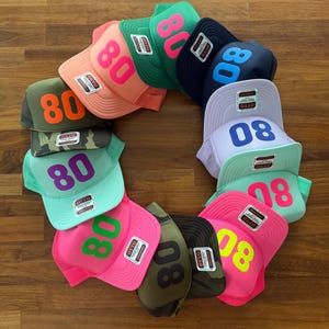 May include: A circular arrangement of multiple Otto brand trucker hats in various colors, including neon pink, green, navy, lavender, and camouflage. Each hat features the number 80 printed on the front panel and a small rectangular brand tag.