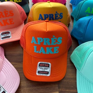 May include: A collection of colorful trucker hats with the text "APR&Egrave;S LAKE" in various colors. The hats have a foam front and mesh back, with a snap closure. The hats are in shades of orange, pink, yellow, green, and blue.