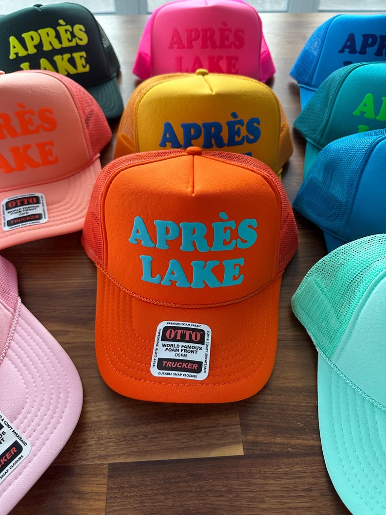 May include: A collection of colorful trucker hats with the text "APR&Egrave;S LAKE" in various colors. The hats have a foam front and mesh back, with a snap closure. The hats are in shades of orange, pink, yellow, green, and blue.
