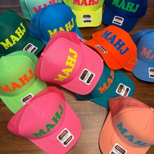 May include: A collection of trucker hats in various colors, including pink, blue, green, and orange. Each hat features the word "MAHJ" in a contrasting color across the front. The hats have a mesh back and a curved brim.