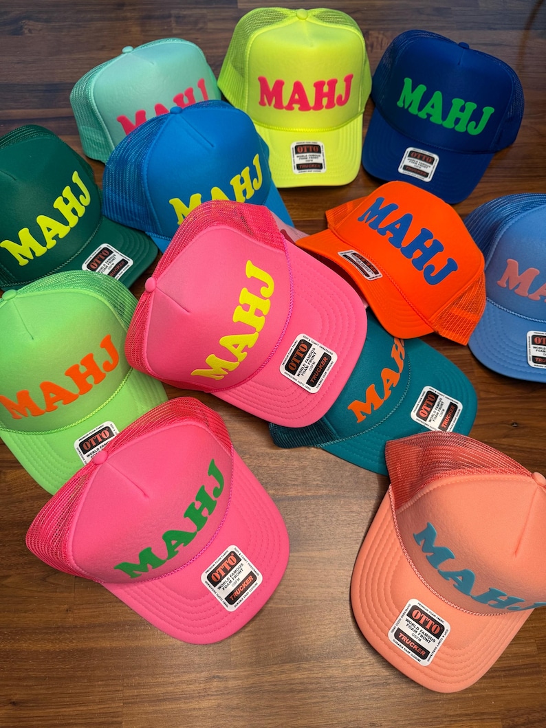 May include: A collection of trucker hats in various colors, including pink, blue, green, and orange. Each hat features the word "MAHJ" in a contrasting color across the front. The hats have a mesh back and a curved brim.
