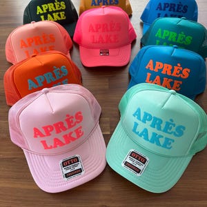 May include: A collection of trucker hats in various colors, including pink, orange, and blue. Each hat features the text "APR&Egrave;S LAKE" in a contrasting color. The hats have a mesh back and a foam front.
