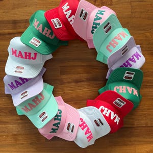 May include: A collection of colorful trucker hats arranged in a circle. The hats are in various colors, including red, green, pink, and white. Each hat features the word "MAHJ" in a contrasting color.