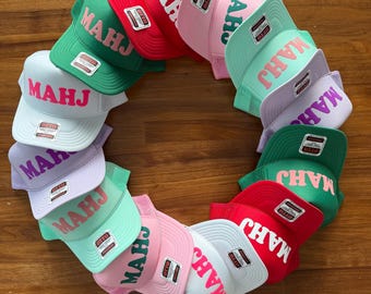May include: A circular arrangement of twelve mesh-back trucker hats in various colors including red, green, light blue, pink, and purple. Each hat features the text MAHJ in bold, contrasting lettering on the front panel and an Otto brand tag.