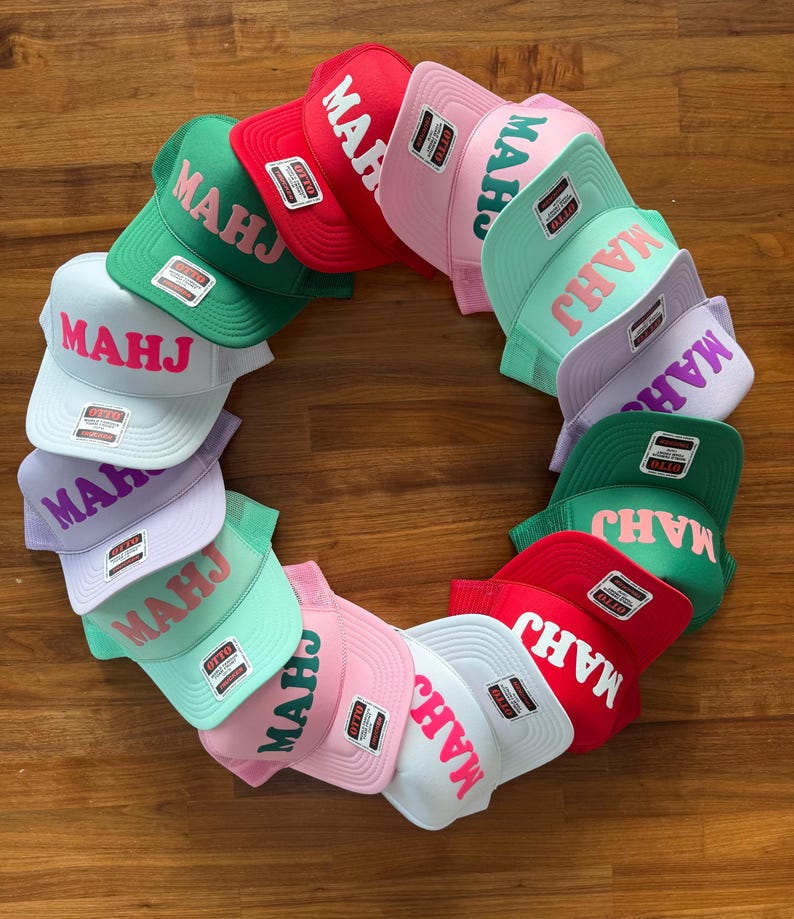 May include: A collection of colorful trucker hats arranged in a circle. The hats are in various colors, including red, green, pink, and white. Each hat features the word "MAHJ" in a contrasting color.