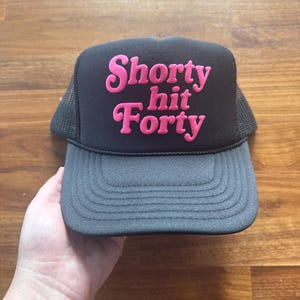 May include: Black and gray trucker hat with the text "Shorty hit Forty" in pink, featuring a mesh back and a curved brim. The hat is held by a hand against a wooden background.