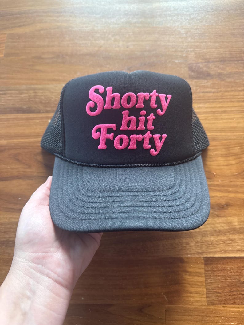 May include: Black and gray trucker hat with the text "Shorty hit Forty" in pink, featuring a mesh back and a curved brim. The hat is held by a hand against a wooden background.