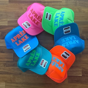 May include: A collection of colorful trucker hats arranged in a circle. Each hat features the text "APR&Egrave;S LAKE" in a contrasting color. The hats are in various colors, including pink, green, blue, and orange.