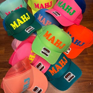 May include: A collection of trucker hats in various colors, including green, pink, orange, and blue. Each hat features the letters "MAHJ" or "CH" in a contrasting color on the front. The hats have a mesh back and a curved brim.