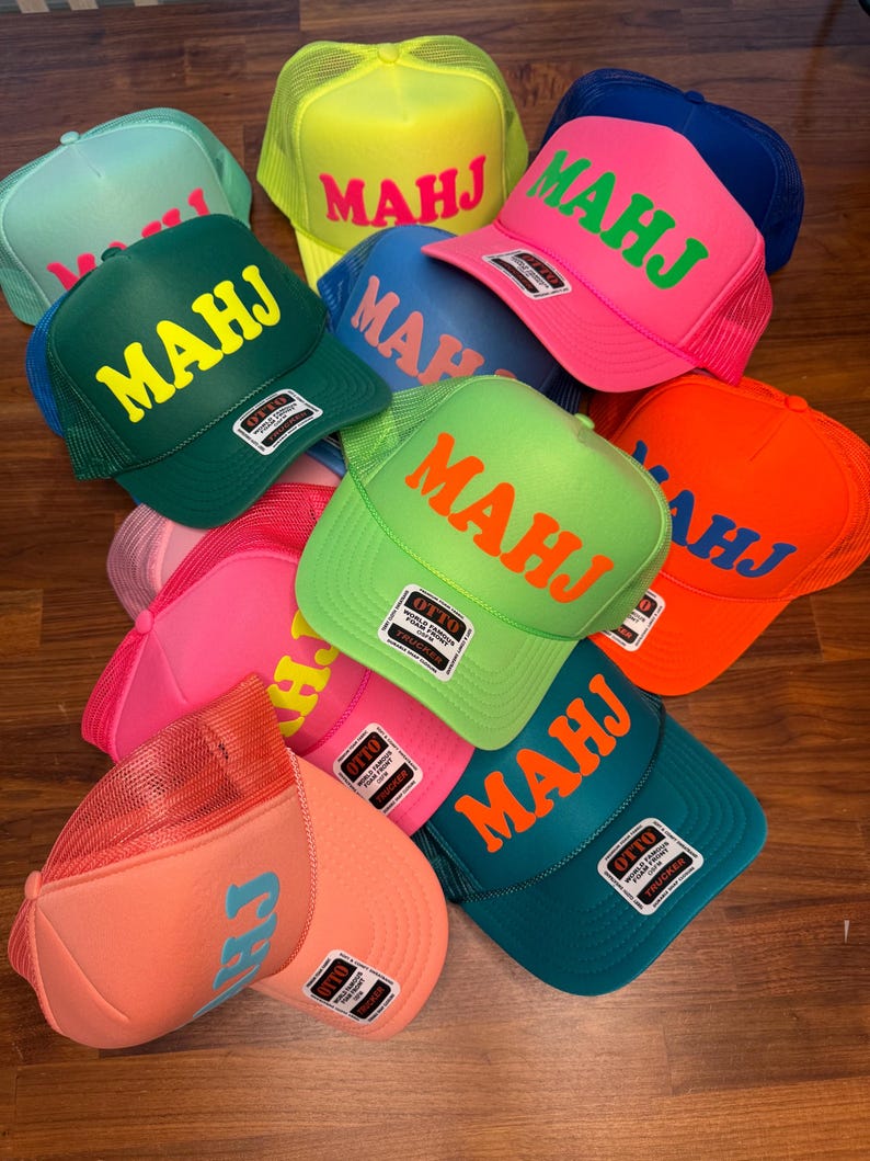May include: A collection of trucker hats in various colors, including green, pink, orange, and blue. Each hat features the letters "MAHJ" or "CH" in a contrasting color on the front. The hats have a mesh back and a curved brim.