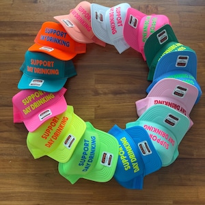 May include: A collection of colorful trucker hats arranged in a circle. Each hat features the text "SUPPORT DAY DRINKING" in a contrasting color. The hats come in various colors, including orange, teal, pink, and green.