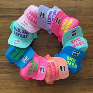 May include: A collection of ten colorful trucker hats arranged in a circle, each featuring unique text such as Pool Please, Poolside Society, Sunshine & Sips, Cabana Club, and It's Pool Time. The hats come in various bright shades including pink, blue, and mint.