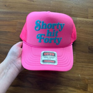 May include: A bright pink trucker hat with the words "Shorty Hit Forty" in teal lettering. The hat has a mesh back and a curved brim. The hat is held by a hand.