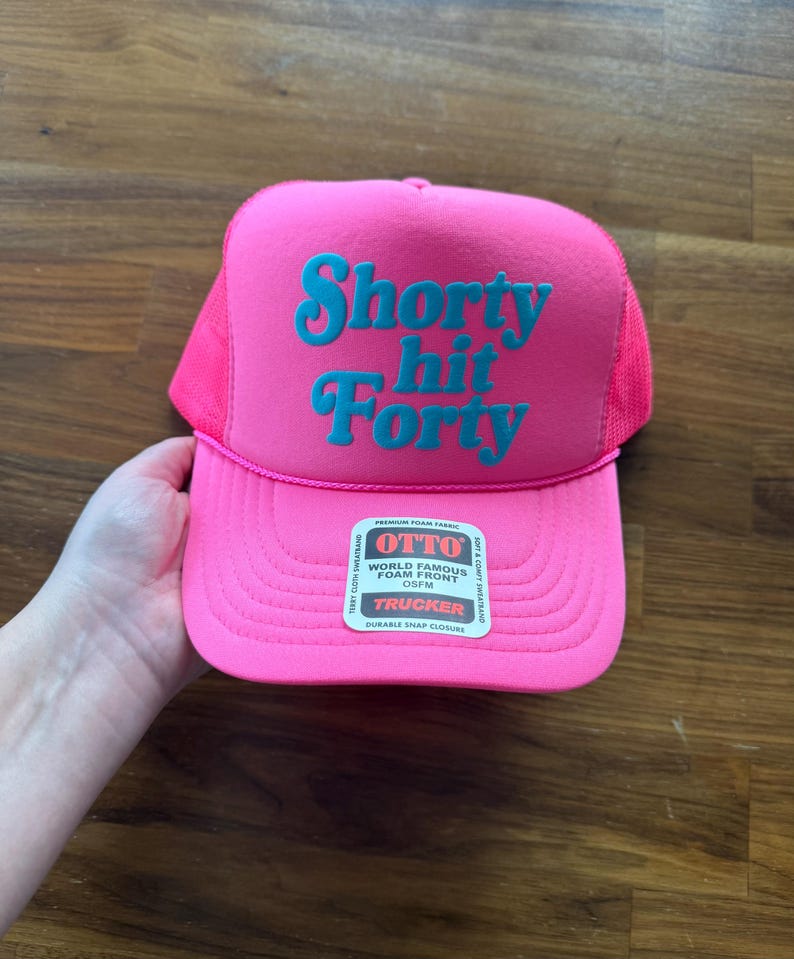 May include: A bright pink trucker hat with the words "Shorty Hit Forty" in teal lettering. The hat has a mesh back and a curved brim. The hat is held by a hand.
