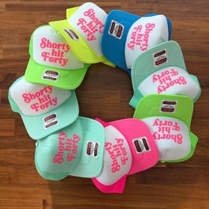 May include: A collection of trucker hats in various colors, including green, blue, pink, and yellow. Each hat features the text "Shorty hit Forty" in pink lettering. The hats are arranged in a circular pattern.