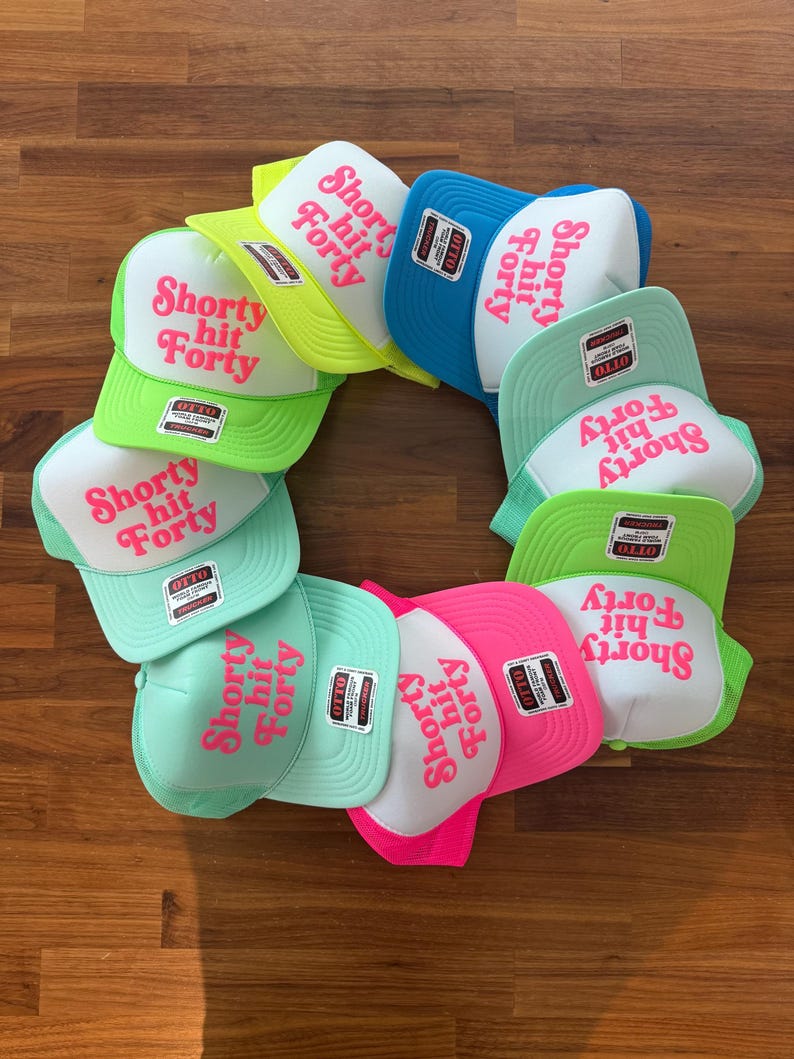 May include: A collection of trucker hats in various colors, including green, blue, pink, and yellow. Each hat features the text "Shorty hit Forty" in pink lettering. The hats are arranged in a circular pattern.