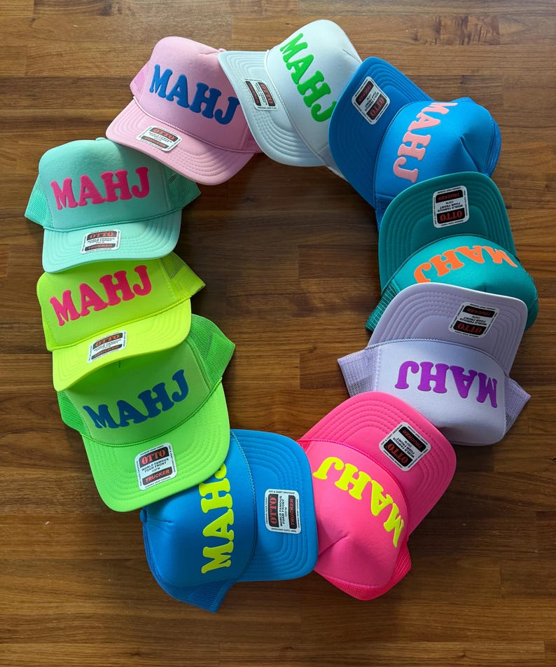 May include: A collection of colorful trucker hats arranged in a circle. Each hat features the word "MAHJ" in a contrasting color. The hats come in various colors, including pink, green, blue, and white, with a mesh back and a flat brim.
