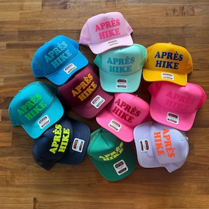May include: A collection of eleven mesh-back trucker hats in various colors, including pink, blue, yellow, green, and purple. Each hat features the text APR&Egrave;S HIKE embroidered on the front panel in a contrasting color and includes an Otto brand tag.