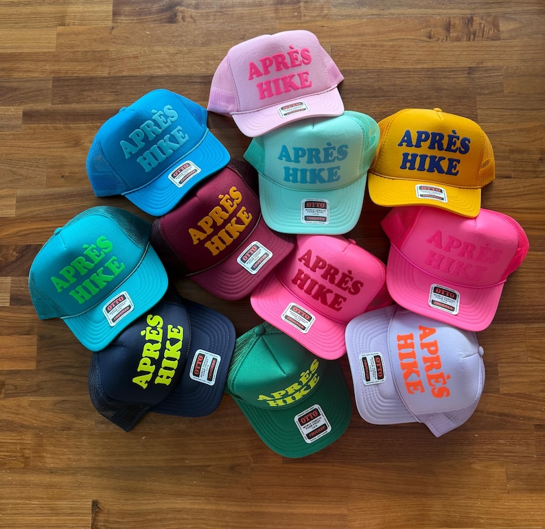 May include: A collection of eleven mesh-back trucker hats in various colors, including pink, blue, yellow, green, and purple. Each hat features the text APR&Egrave;S HIKE embroidered on the front panel in a contrasting color and includes an Otto brand tag.