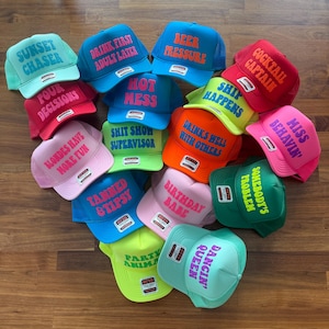 May include: A collection of colorful foam trucker hats featuring various phrases in bold lettering, including Sunset Chaser, Drink First Adult Later, Beer Pressure, Cocktail Captain, Four Decisions, Hot Mess, Ship Happens, Miss Behavin, and Party Animal.