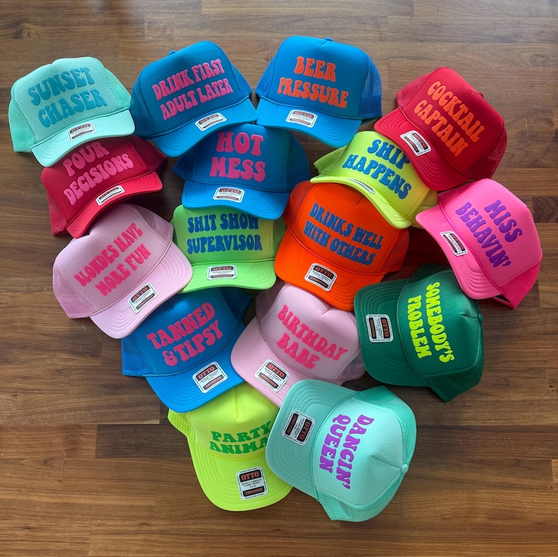 May include: A collection of colorful foam trucker hats featuring various phrases in bold lettering, including Sunset Chaser, Drink First Adult Later, Beer Pressure, Cocktail Captain, Four Decisions, Hot Mess, Ship Happens, Miss Behavin, and Party Animal.