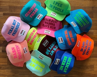 May include: A collection of twelve colorful foam trucker hats with mesh backs, each featuring unique text printed on the front. The hats come in various bright colors including pink, blue, green, orange, and yellow, with contrasting text colors.