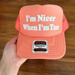 May include: A coral-colored trucker hat featuring a mesh back and a foam front panel. The text "I'm Nicer When I'm Tan" is printed in white on the front. A rectangular sticker on the brim reads "OTTO WORLD FAMOUS FOAM FRONT OSFM TRUCKER".