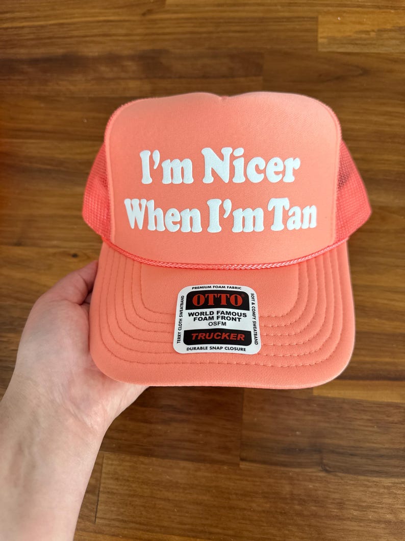 May include: A coral-colored trucker hat featuring a mesh back and a foam front panel. The text "I'm Nicer When I'm Tan" is printed in white on the front. A rectangular sticker on the brim reads "OTTO WORLD FAMOUS FOAM FRONT OSFM TRUCKER".