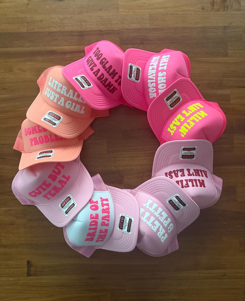 May include: A circular arrangement of ten trucker hats in various shades of pink and orange. Each hat features an OTTO brand sticker and white or yellow text including phrases like LITERALLY JUST A GIRL, TOO GLAM TO GIVE A DAMN, and MILFIN AIN'T EASY.
