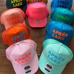 May include: A collection of trucker hats in various colors, including green, yellow, pink, blue, and orange. Each hat features the text "APR&Egrave;S LAKE" in a contrasting color. The hats have a mesh back and a curved brim.