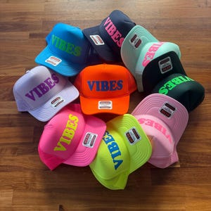 May include: A collection of ten foam trucker hats in various bright colors, including neon pink, orange, lime green, light blue, and lavender. Each hat features the word VIBES printed in a contrasting color on the front panel and an Otto brand tag.