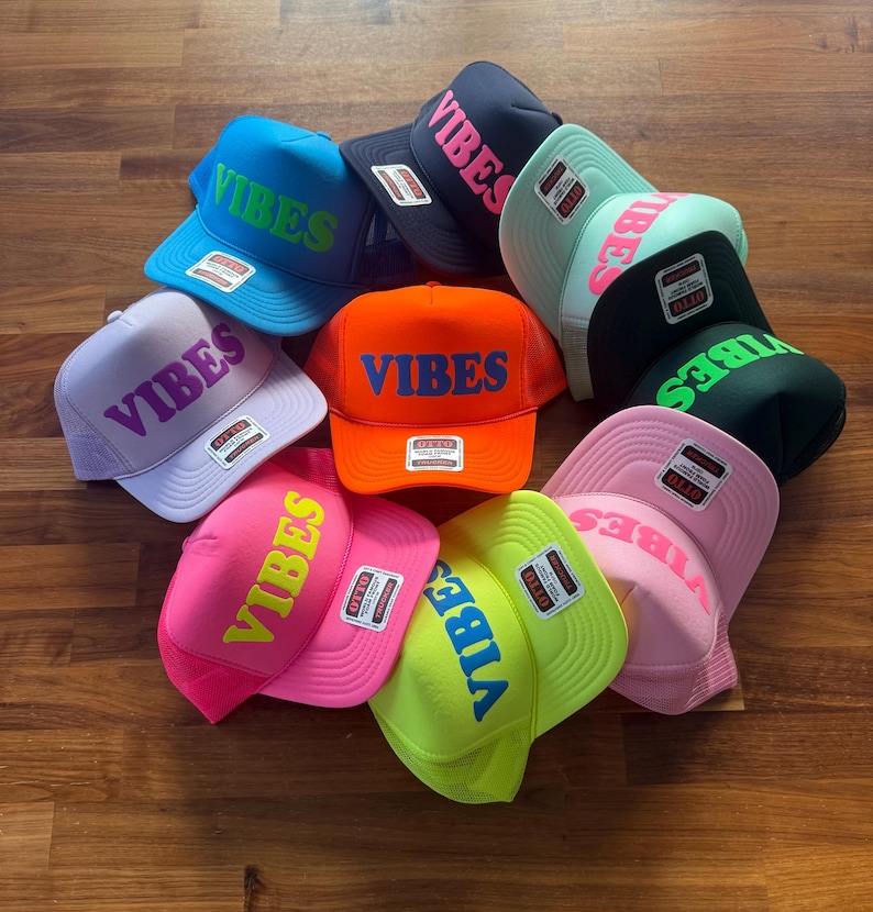 May include: A collection of ten foam trucker hats in various bright colors, including neon pink, orange, lime green, light blue, and lavender. Each hat features the word VIBES printed in a contrasting color on the front panel and an Otto brand tag.