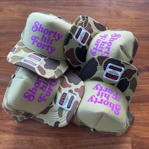 May include: A collection of trucker hats with a camouflage pattern on the brim and a tan front panel. The hats feature the text "Shorty Hit Forty" in bright purple. Each hat has a mesh back and a small tag.