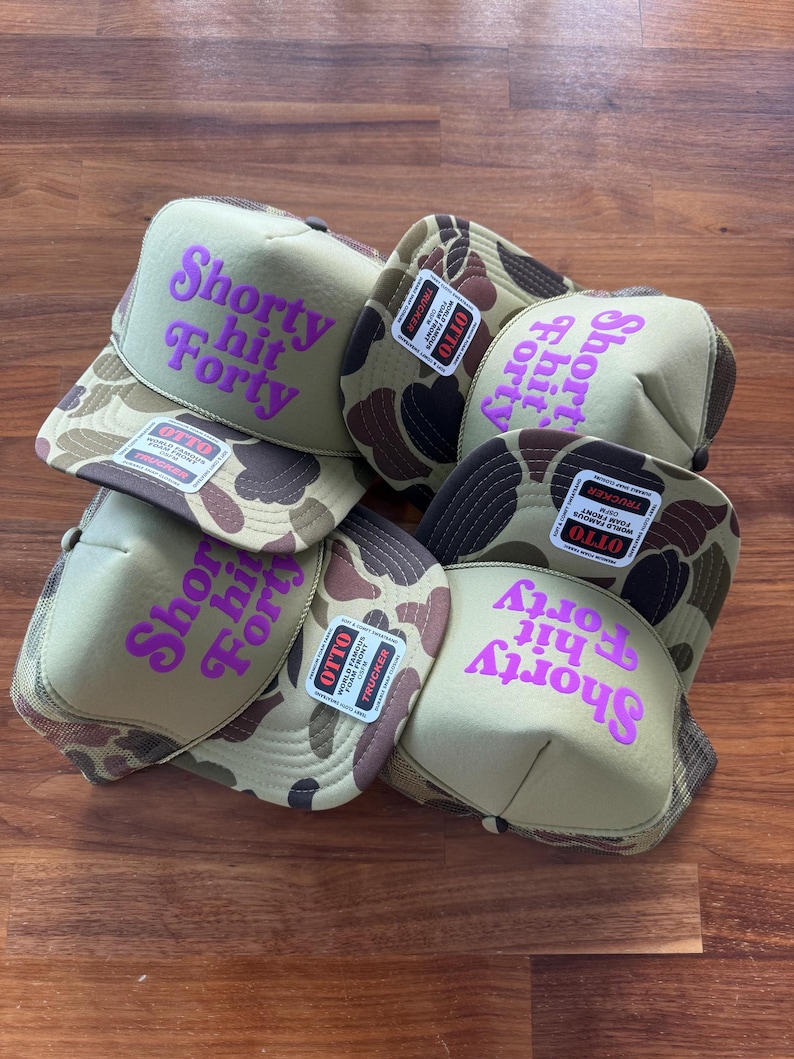 May include: A collection of trucker hats with a camouflage pattern on the brim and a tan front panel. The hats feature the text "Shorty Hit Forty" in bright purple. Each hat has a mesh back and a small tag.