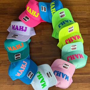 May include: A collection of colorful trucker hats arranged in a circle. Each hat features the word "MAHJ" in a contrasting color across the front. The hats come in various colors, including pink, blue, green, and white.