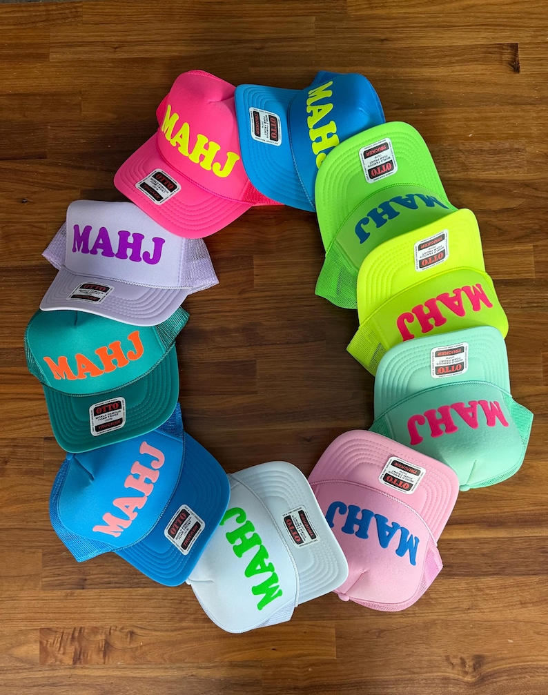 May include: A collection of colorful trucker hats arranged in a circle. Each hat features the word "MAHJ" in a contrasting color across the front. The hats come in various colors, including pink, blue, green, and white.
