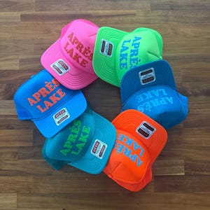 May include: A collection of trucker hats in a circular arrangement. The hats are in various bright colors, including pink, green, blue, and orange. Each hat features the text "APR&Egrave;S LAKE" in a contrasting color on the front.