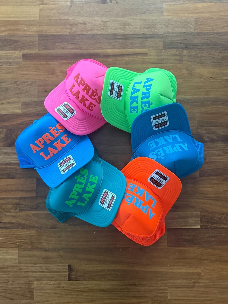 May include: A collection of trucker hats in a circular arrangement. The hats are in various bright colors, including pink, green, blue, and orange. Each hat features the text "APR&Egrave;S LAKE" in a contrasting color on the front.