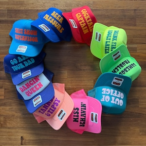 May include: A circular arrangement of twelve colorful trucker hats featuring various slogans including SHIT SHOW SUPERVISOR, BEER PRESSURE, COCKTAIL CAPTAIN, DRINK FIRST ADULT LATER, SOMEBODY ELSE'S PROBLEM, OUT OF OFFICE, MISS BEHAVIN', TANNER & TIPSY, DANCIN' QUEEN, and GO ASK YOUR DAD.