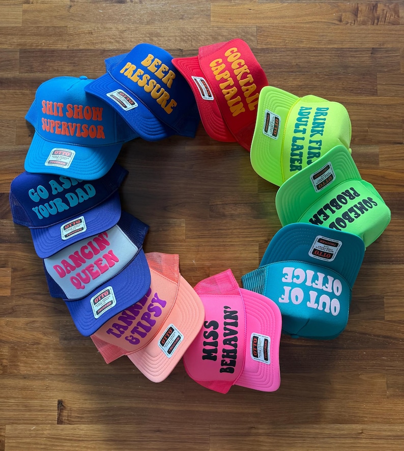May include: A circular arrangement of twelve colorful trucker hats featuring various slogans including SHIT SHOW SUPERVISOR, BEER PRESSURE, COCKTAIL CAPTAIN, DRINK FIRST ADULT LATER, SOMEBODY ELSE'S PROBLEM, OUT OF OFFICE, MISS BEHAVIN', TANNER & TIPSY, DANCIN' QUEEN, and GO ASK YOUR DAD.