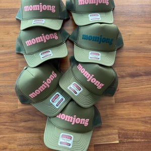May include: A collection of seven olive green trucker hats with mesh backs and foam fronts. Each hat features the text momjong embroidered in either pink or dark blue on the front panel and includes an Otto brand sticker on the brim.