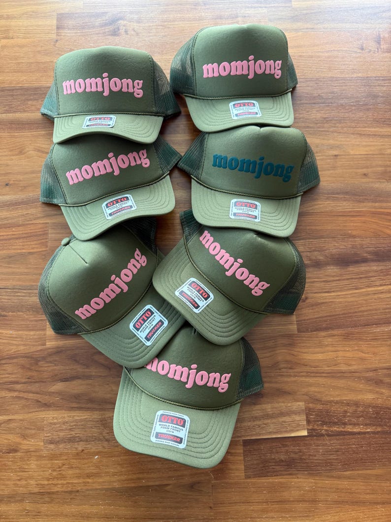 May include: A collection of seven olive green trucker hats with mesh backs and foam fronts. Each hat features the text momjong embroidered in either pink or dark blue on the front panel and includes an Otto brand sticker on the brim.