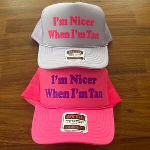 May include: Two trucker hats with mesh backs and foam fronts. The top hat is light purple with hot pink text reading I'm Nicer When I'm Tan. The bottom hat is bright pink with purple text reading I'm Nicer When I'm Tan. Both have Otto brand labels.