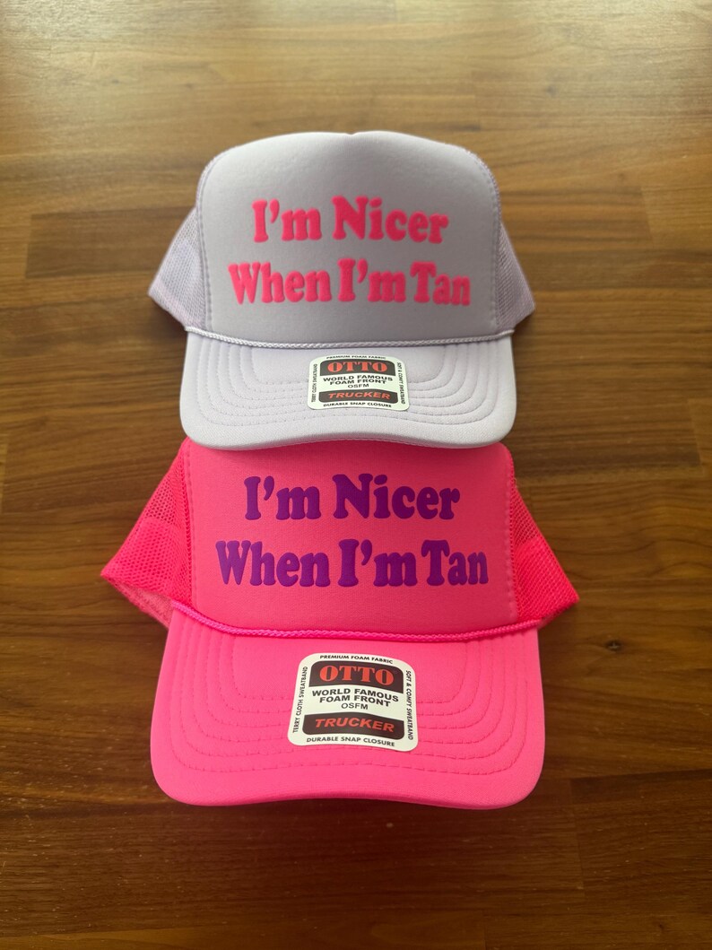May include: Two trucker hats with mesh backs and foam fronts. The top hat is light purple with hot pink text reading I'm Nicer When I'm Tan. The bottom hat is bright pink with purple text reading I'm Nicer When I'm Tan. Both have Otto brand labels.